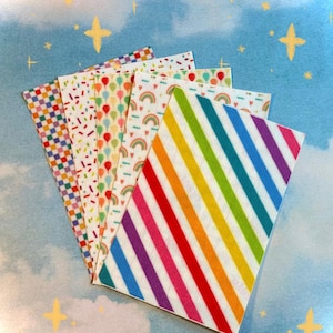 Könnte beinhalten: Eine Sammlung farbenfroher Papiere mit Mustern, die vor einem blauen Hintergrund mit goldenen Sternen aufgefächert sind. Die Papiere zeigen Designs wie Regenbogenstreifen, Ballons, Regenbogen und Schachbrettmuster. Das Gesamtthema ist hell und fröhlich.