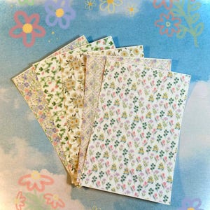 Könnte beinhalten: Eine Sammlung von Papierbögen mit floralen Mustern. Die Bögen zeigen Muster aus kleinen gelben, lila und grünen Blumen auf weißem Hintergrund. Der Hintergrund ist hellblau mit gezeichneten Blumen.
