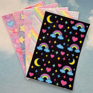 Könnte beinhalten: Eine Sammlung bunter Aufkleber mit verschiedenen Designs. Die Aufkleber zeigen Herzen, Sterne, Regenbogen und Monde auf schwarzem Hintergrund. Andere Aufkleber haben Pastellmuster von Schleifen, Sternen und Süßigkeiten.