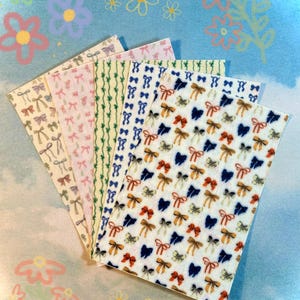 May include: A collection of patterned stickers featuring various designs, including bows and vertical lines. The stickers display a range of colors, such as beige, pink, green, and blue, against a white background. The background has a light blue watercolor effect with flower illustrations.