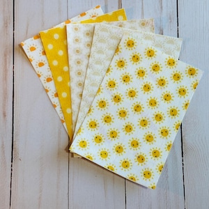 May include: Six rectangular fabric pieces in a fan arrangement, featuring yellow and white patterns. Designs include polka dots, smiling suns, and sunbursts. The fabrics are displayed on a light wood surface.