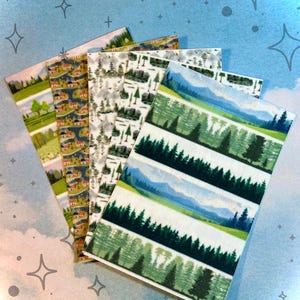 Könnte beinhalten: Eine Sammlung dekorativer Washi-Tapes mit Landschaftsdesigns. Die Tapes zeigen Szenen von Wäldern, Bergen und ländlichen Umgebungen mit Häusern und Bäumen. Die Farbpalette umfasst Grün-, Blau- und Erdtöne.
