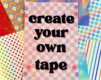 Puede incluir: Una colección de cintas decorativas coloridas. La cinta central presenta el texto "create your own tape" en letras negras sobre un fondo a cuadros rosa y blanco. Otras cintas tienen patrones de arcoíris, estrellas y lunares.