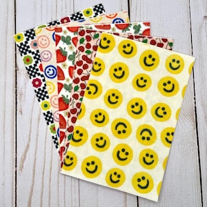 Könnte beinhalten: Eine Sammlung farbenfroher, gemusterter Tücher. Designs umfassen schwarz-weißes Schachbrettmuster, Smiley-Gesichter, Erdbeeren, Kirschen und Blumen. Die Tücher sind fächerförmig vor einem hellen Holzmaserungshintergrund angeordnet.