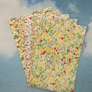 Op de afbeelding: Een waaier van vijf vellen papier met bloemmotieven. De patronen omvatten gele, rode, blauwe en witte bloemen met groene bladeren. De achtergrond is lichtblauw met witte wolken.