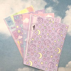 Puede incluir: Un abanico de pegatinas de colores pastel con varios dise&ntilde;os. Las pegatinas presentan osos de peluche, lazos, nubes, estrellas y lunas. La paleta de colores incluye rosa, morado, amarillo y azul, creando una est&eacute;tica caprichosa y linda.