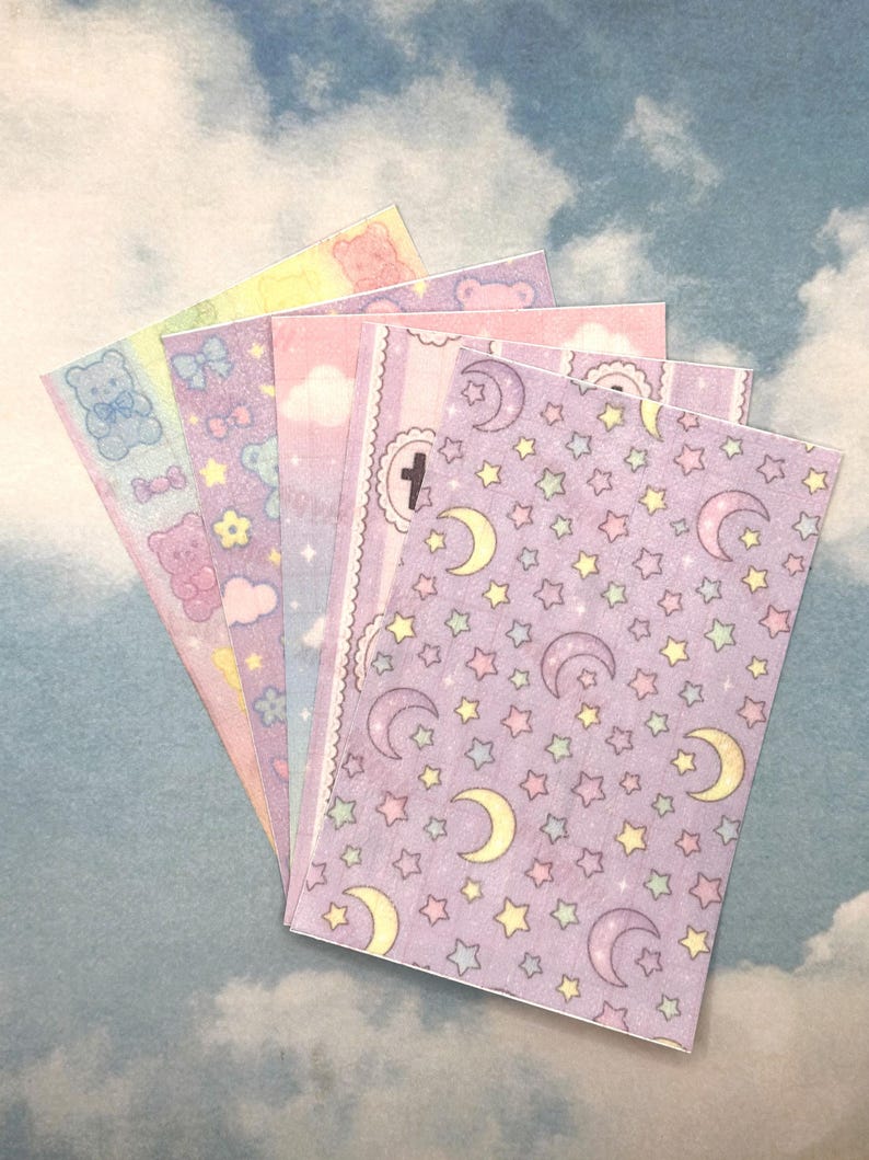 Puede incluir: Un abanico de pegatinas de colores pastel con varios dise&ntilde;os. Las pegatinas presentan osos de peluche, lazos, nubes, estrellas y lunas. La paleta de colores incluye rosa, morado, amarillo y azul, creando una est&eacute;tica caprichosa y linda.