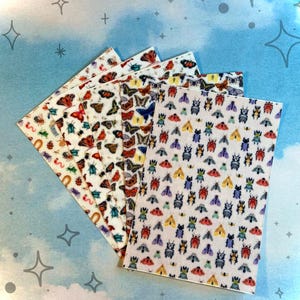 Puede incluir: Una colección de pegatinas rectangulares con diseños de insectos variados, incluyendo mariposas, polillas y escarabajos. Las pegatinas muestran una gama de colores y patrones, con un fondo blanco. Las pegatinas están dispuestas en forma de abanico sobre un fondo azul.