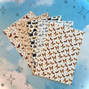 Puede incluir: Una colección de hojas de papel estampadas con diversos diseños de mariposas y polillas. Las hojas muestran una gama de colores, incluyendo marrón, beige, negro y azul, con ilustraciones detalladas de insectos. El fondo es azul claro con formas de estrellas y cruces.