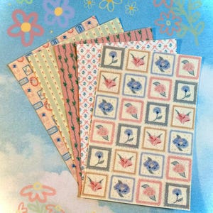 Puede incluir: Una colección de hojas de papel estampadas en colores pastel, incluyendo rosa, verde y crema. Los diseños presentan patrones florales y geométricos, con un patrón cuadrado de flores en primer plano. El fondo es azul claro con ilustraciones de flores.