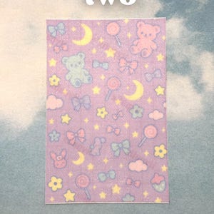 Puede incluir: Adhesivo rectangular con un patr&oacute;n repetitivo de ositos de peluche, lazos, piruletas, estrellas, lunas y flores sobre un fondo lavanda. La palabra "two" est&aacute; impresa en blanco en la parte superior.