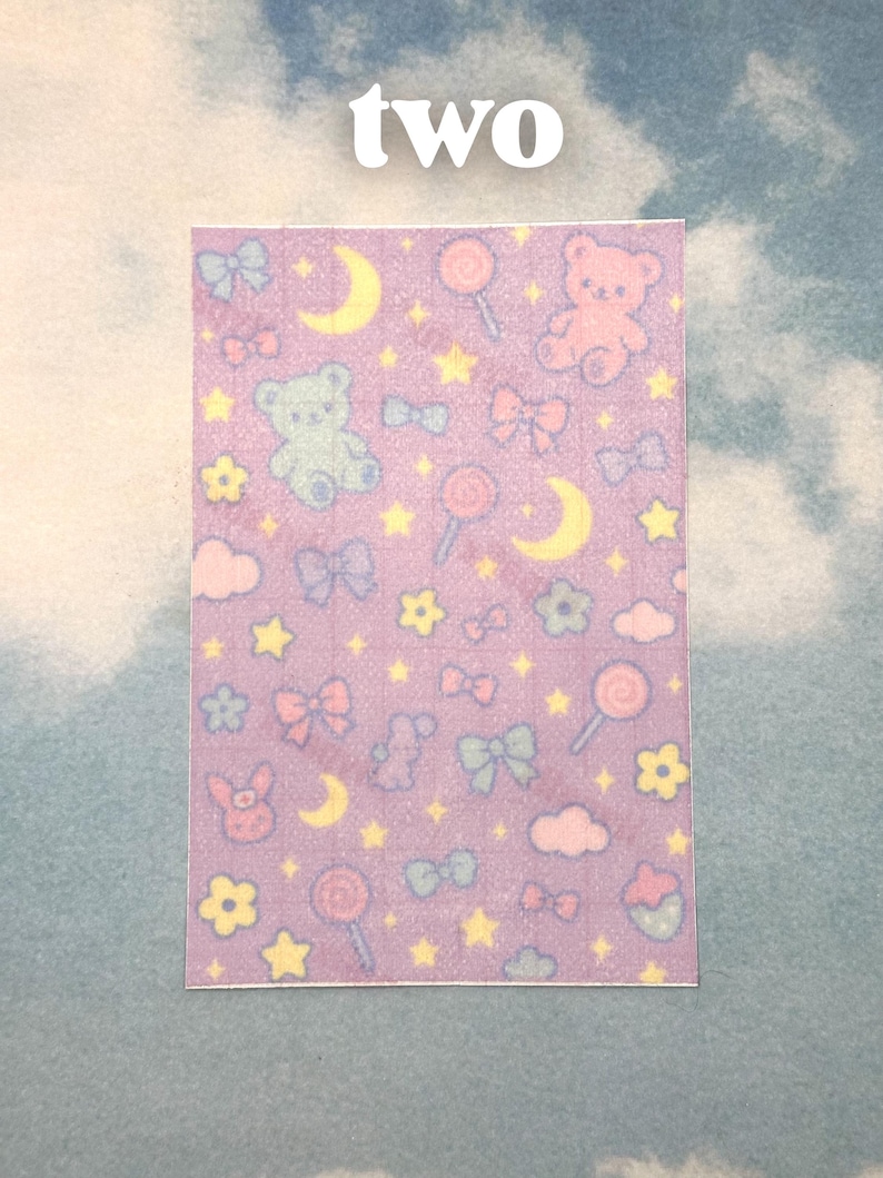 Puede incluir: Adhesivo rectangular con un patr&oacute;n repetitivo de ositos de peluche, lazos, piruletas, estrellas, lunas y flores sobre un fondo lavanda. La palabra "two" est&aacute; impresa en blanco en la parte superior.