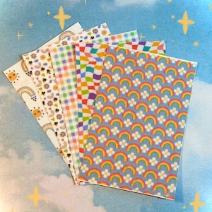 Könnte beinhalten: Eine Sammlung von Papieren mit verschiedenen Mustern, darunter Regenbogen, Bienen, Blumen und Karos. Die Farbpalette umfasst Pastelltöne und helle Farben, mit einem blauen Himmel Hintergrund und Sternen.