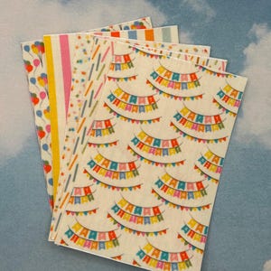 Könnte beinhalten: Ein Stapel dekorativer Papierbögen mit bunten Mustern. Designs umfassen Ballons, Streifen, Konfetti und sich wiederholende Reihen festlicher Wimpelketten. Die Papiere sind vor einem hellblauen Hintergrund angeordnet.
