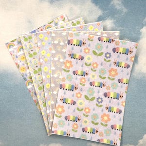 Könnte beinhalten: Eine Sammlung von Papieren mit Pastellmustern. Die Muster umfassen Regenbogen, lächelnde Sonnen, Bäume und bunte Raupen mit Blumen. Die Papiere sind fächerförmig vor einem Hintergrund mit bewölktem Himmel angeordnet.