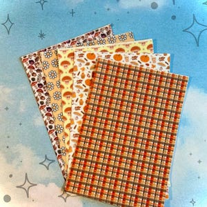 May include: A stack of patterned paper sheets with autumn-themed designs. The patterns include mushrooms, daisies, pumpkins, and plaid in shades of brown, orange, and yellow. The background is a light blue with star and cross shapes.