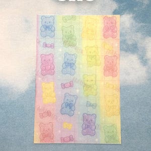 Puede incluir: Una hoja de pegatinas con un fondo degradado de arco&iacute;ris. Las pegatinas presentan ositos de peluche brillantes en varios colores, cada uno con una pajarita. La palabra "one" est&aacute; escrita en blanco en la parte superior.