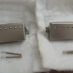 Può includere: Due pickup humbucker argentati per chitarra, ciascuno con sei viti visibili, e due set di molle argentate. I pickup rettangolari hanno una finitura in metallo spazzolato. Gli oggetti sono su una superficie bianca strutturata.