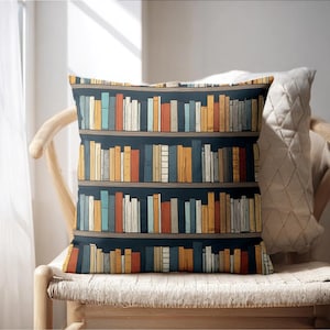 May include: Decorative throw pillow featuring a bookshelf design. The pillow displays a dark blue background with three shelves filled with colorful books in shades of orange, yellow, blue, and red. The pillow is placed on a wicker chair.