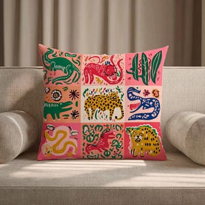 May include: A square decorative pillow with a pink background and a grid pattern of animal illustrations. The illustrations include a green dinosaur, a red cat, a yellow leopard, and a blue snake. The pillow is on a gray sofa.