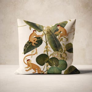 Puede incluir: Cojín cuadrado con fondo crema, que presenta una ilustración botánica de monos entre hojas y enredaderas verdes. Los monos son marrones con gorras rojas. El cojín tiene un aspecto suave, adecuado para la decoración del hogar.