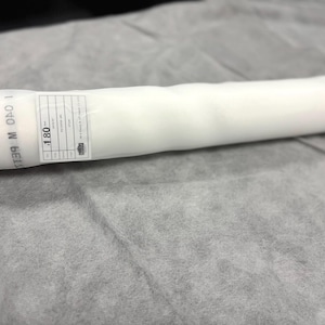 May include: A rolled white fabric, possibly a filter or material for a larger project. The roll is wrapped in clear plastic, with a label showing text and measurements. The fabric is lying on a gray surface.