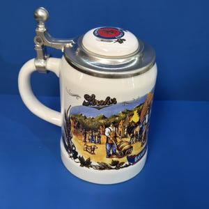 May include: White ceramic beer stein with a hinged metal lid and handle. The stein features a colorful illustration of a Bavarian village scene. The lid has a red and blue emblem. The word "Bavaria" is visible on the side.
