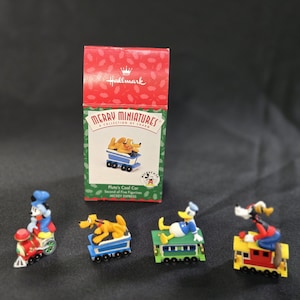 May include: A collection of Hallmark Merry Miniatures featuring Disney characters. The set includes Pluto's Coal Car and other figurines. The box is red with the Hallmark logo. The figurines are on a black surface.
