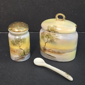 May include: A porcelain salt shaker with a gold-tone lid, a matching lidded jar with a gold-tone handle, and a small spoon. Both containers feature a hand-painted landscape scene with trees and a yellow sky against a white background.