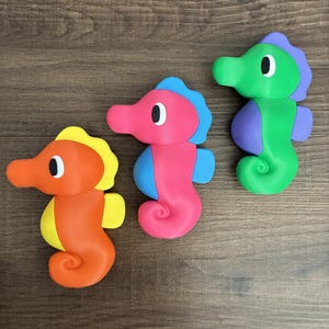 May include: Three colorful seahorse-shaped toys in orange, pink, and green. Each has a contrasting color for the fin and head crest. The toys are arranged on a brown wooden surface.