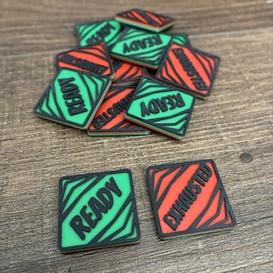 May include: A collection of square, two-tone, decorative tiles. The tiles are green and red with black borders. The word "READY" is printed on some tiles, and "EXHAUSTED" on others. The tiles are scattered on a wooden surface.