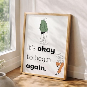 Può includere: Una stampa artistica incorniciata con una cornice in legno chiaro. La stampa presenta illustrazioni a tratteggio di due figure e il testo "It's okay to begin again." Le figure indossano una felpa con cappuccio verde e pantaloni arancioni.