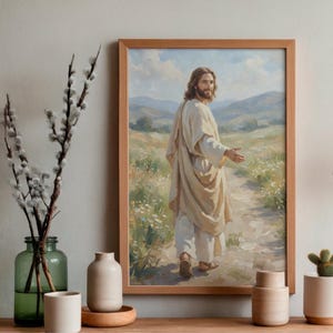 May include: A framed print of a painting depicting a figure in a cream-colored robe walking on a path. The artwork features a light brown wooden frame and a serene landscape with mountains and a cloudy sky.