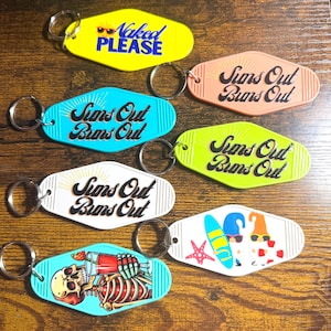 Könnte beinhalten: Eine Sammlung farbenfroher Schlüsselanhänger im Motel-Stil. Die Schlüsselanhänger zeigen verschiedene Designs und Texte, darunter "Naked PLEASE" und "Suns Out Buns Out". Weitere Designs sind ein Skelett, das einen Cocktail trinkt, Surfbretter und Zwerge.