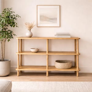 May include: A light-colored wooden console table with three shelves. The table has a natural wood finish and is supported by cylindrical legs. A woven basket, a ceramic bowl, and a stack of books are on the shelves. A framed landscape painting hangs above the table.