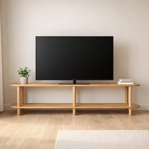 May include: A light-colored wooden TV stand with a large black screen television on top. The stand has two shelves and rounded edges. A small potted plant and books are on the stand. The stand is on a light wood floor.