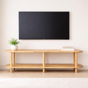 May include: A light-colored wooden TV stand with an oval top and a lower shelf. The stand has four cylindrical legs and supports a large, black flat-screen TV. A small potted plant and books are on the stand.