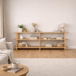 May include: A light-colored wooden bookshelf with three shelves, holding books, decorative items, and plants. The shelves are supported by vertical posts. A lamp and a sofa are visible in the background, creating a cozy living room setting.