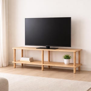 May include: A light-colored wooden TV stand with two shelves and rounded legs. A large black television sits on top. A small potted plant and a stack of books are on the lower shelf. The stand is on a light-colored wooden floor.