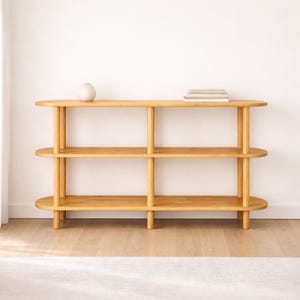 May include: A light-colored wooden bookshelf with three shelves and vertical supports. The shelves are oval-shaped, and the unit is empty except for a small vase and a stack of books. The bookshelf is against a white wall.