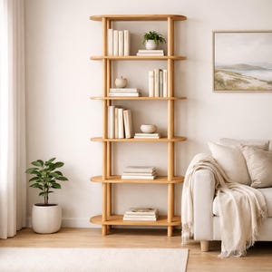 May include: A light oak bookshelf with five shelves, holding books, small plants, and decorative items. The shelves have rounded edges and are supported by vertical wooden posts. The bookshelf is in a well-lit room with a white wall and a sofa.