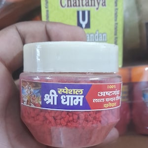 May include: A small, clear jar filled with red granules, labeled in Hindi with the words "Shri Dham" and "Special." The jar has a white lid and is held in a person's hand. The background includes a box with text.