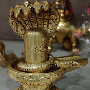 Könnte beinhalten: Eine goldene Shiva Lingam Statue aus Messing. Die Skulptur zeigt eine zentrale Säule mit einer Schlange, die sich darum windet, und eine Kobrakappe darüber. Der Sockel ist aufwendig mit Mustern verziert. Eine weitere goldene Statue befindet sich im Hintergrund.