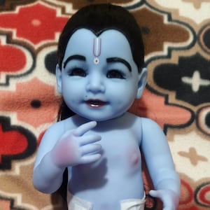 Könnte beinhalten: Eine blauhäutige Babypuppe mit schwarzen Haaren und einer weißen Windel. Die Puppe hat ein bemaltes Gesicht mit dunklen Augen und roter Lippenfarbe. Die Puppe sitzt auf einer gemusterten Decke.