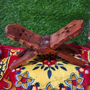 Puede incluir: Un atril de madera marrón oscuro, diseñado para sostener un libro abierto, se muestra sobre un textil estampado en rojo y amarillo. El atril tiene un diseño en forma de cruz. El textil presenta patrones florales y geométricos.