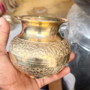 Puede incluir: Una pequeña y ornamentada olla de latón con un borde ancho y patrones florales detallados. La olla se sostiene en una mano, mostrando su tamaño y artesanía. La superficie de la olla refleja la luz.