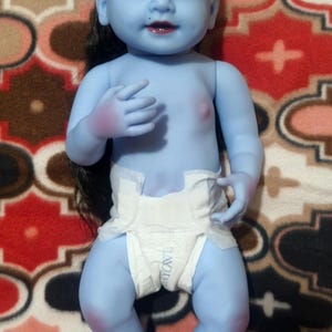 May include: A blue-skinned baby doll with black hair and a white diaper. The doll has a painted face with dark eyes, a red mouth, and a purple mark on its forehead. The doll is lying on a patterned red and brown surface.
