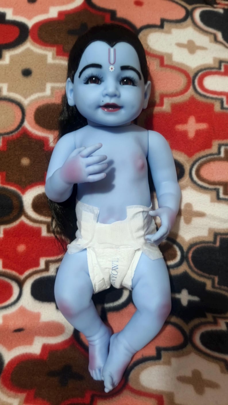 May include: A blue-skinned baby doll with black hair and a white diaper. The doll has a painted face with dark eyes, a red mouth, and a purple mark on its forehead. The doll is lying on a patterned red and brown surface.
