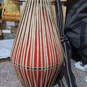 Puede incluir: Un instrumento de percusión tradicional indio, el mridangam, con un cuerpo rojo y un marco de bambú tejido. Una correa azul y blanca está unida al instrumento.