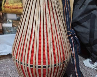 May include: A traditional Indian percussion instrument, the mridangam, with a red body and a woven bamboo frame. A blue and white strap is attached to the instrument.